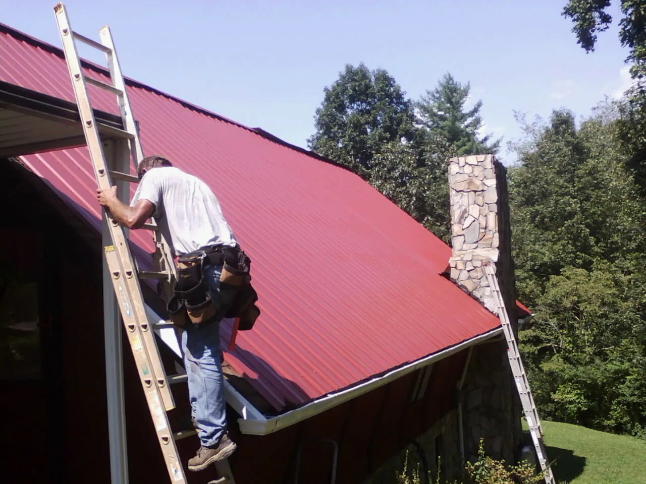 Roofer on a ladder beside a red metal roof and stone chimney — Soffit & Fascia Repair by Roofing Brothers in Hempstead